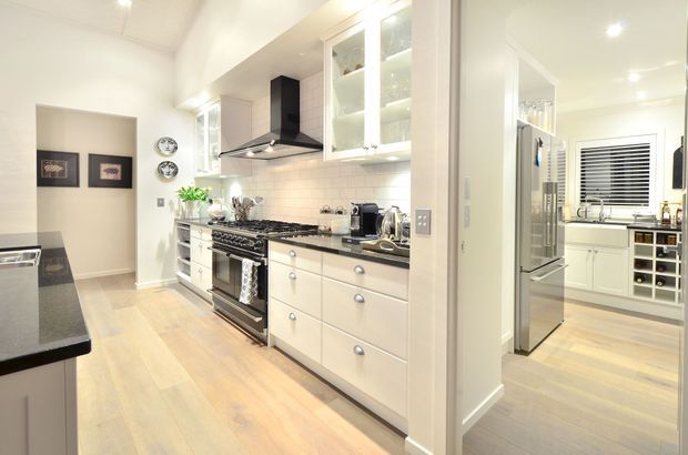 Bright, spacious white kitchen with pale timber flooring, white shaker-style cabinetry, dark stone benchtops, a tiled splashback, stainless-steel range cooker and black rangehood. Glazed upper cabinets, open shelving, and a large opening provide a view into an adjoining utility or pantry area with additional cabinetry and a wine rack.