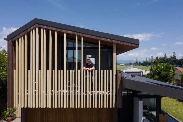 Exterior view of a contemporary multi-level residential building with a sharply projecting flat roof and a screened balcony or upper-level facade. Vertical timber battens create a strong privacy screen over dark timber cladding and extensive glazing. The building sits in a suburban setting with distant mountains and water visible beyond, under a clear blue sky.