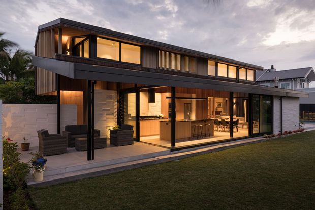 Exterior view of a sophisticated two-storey contemporary residence at dusk. The home features a dark, rectilinear facade, extensive floor-to-ceiling glazing, a broad covered terrace, vertical timber screening, warm interior lighting, and an open-plan kitchen and dining area visible through the glass. A landscaped lawn and paved outdoor entertaining area sit in front of the house.