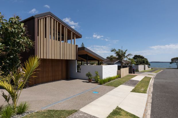 Contemporary two-storey coastal residence viewed from the street, featuring a dark timber-clad lower level, an upper screen of closely spaced vertical timber battens, a broad flat roof, and a covered carport or entry canopy. White rendered boundary walls, concrete driveway and footpath, tropical landscaping, and glimpses of the waterfront contribute to the setting.