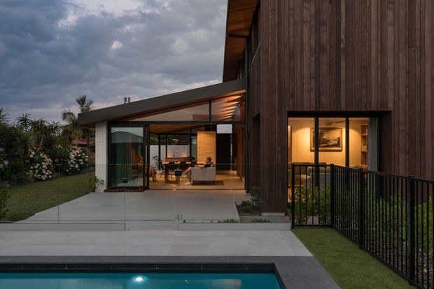 Dusk exterior view of a contemporary multi-level residence, combining dark vertical timber cladding with a low-slung glazed living pavilion. A paved terrace and swimming pool occupy the foreground, while warm interior lighting reveals the living and dining areas through floor-to-ceiling glazing. Black metal balustrading, landscaped planting, and a covered outdoor threshold complete the scene beneath a dramatic cloudy sky.