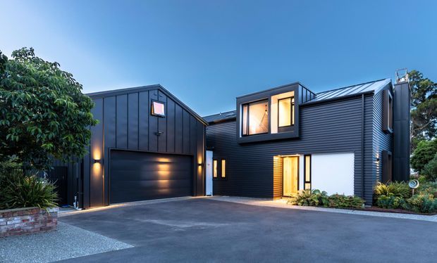A contemporary dark-clad residential house viewed from the driveway at dusk. The two-storey home features vertically and horizontally profiled dark metal cladding, a prominent double garage, angular pitched roof forms, large illuminated windows, and a recessed timber-toned entry door. Low-maintenance landscaping and paved areas frame the facade.