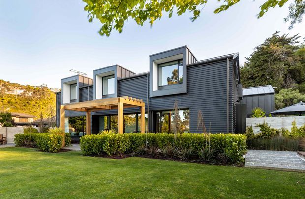 A contemporary multi-level home viewed from the landscaped front garden. The dark horizontal-clad facade features projecting white-trimmed window bays, a timber pergola over an outdoor terrace, large glazed openings, and a low-pitched roofline. Neatly clipped hedges, lawn, planting beds, and a paved path frame the house.