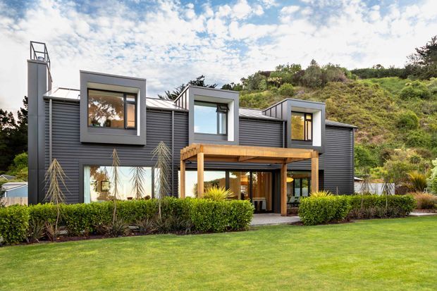 Modern two-storey residential house set against a vegetated hillside, with dark horizontal exterior cladding, projecting box-framed windows, a flat roofline, and a timber-covered outdoor terrace. The landscaped foreground includes a manicured lawn and clipped hedges.