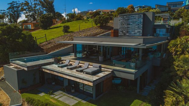 A contemporary multi-level hillside residence with extensive floor-to-ceiling glazing, broad projecting roof planes, a rooftop terrace, and a rectangular swimming pool. The house is integrated into a landscaped sloping site, with an elevated pool deck, outdoor lounge furniture, and a patterned feature wall on the upper level.