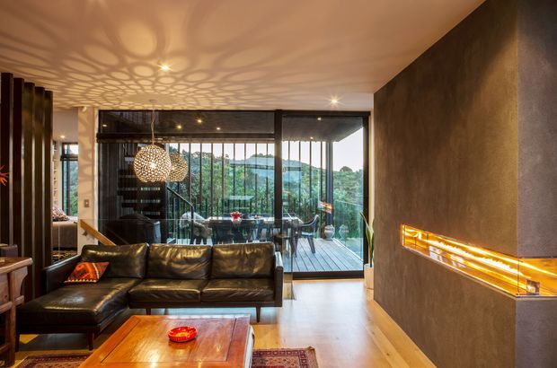 A warm, sophisticated living room with a dark leather sectional and timber coffee table, centred around a long built-in linear fireplace set into a textured dark feature wall. Floor-to-ceiling glazing opens to a timber balcony and leafy hills beyond. Decorative pendant lights cast patterned shadows across the ceiling, while dark vertical screens and glazing frames add architectural definition.