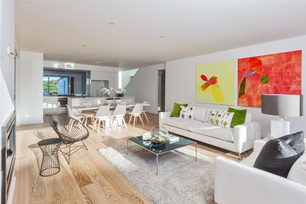 Bright open-plan contemporary living and dining area with pale timber flooring, white walls and furnishings, a white sofa arrangement, glass coffee table, sculptural wire chairs, and colourful abstract artwork. The dining area and kitchen are visible beyond, with abundant daylight and a clean, spacious layout.