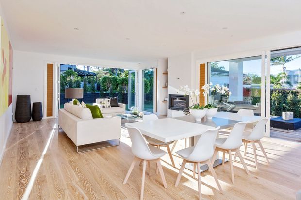 Bright open-plan contemporary living and dining area with white walls, pale timber flooring, extensive floor-to-ceiling glazing, and doors opening to a landscaped outdoor area. A white sectional sofa, white dining table, sculptural white chairs, indoor plants, and a compact fireplace create a clean, airy interior.