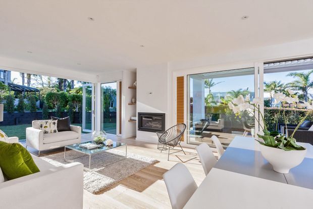 Bright open-plan living and dining area with floor-to-ceiling glazing, white walls and ceiling, pale timber flooring, a minimalist built-in fireplace, white dining table and chairs, and contemporary lounge furniture. Lush garden planting and a covered outdoor area are visible through the glazing.