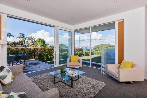 Bright contemporary living room with floor-to-ceiling corner glazing and sliding glass doors opening to a timber deck and garden. The room contains two light upholstered sofas, a glass coffee table, a textured rug, and yellow accent cushions, with expansive views of greenery and water beyond.