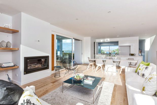 Bright, open-plan contemporary living and dining area with a white palette, pale timber flooring, a built-in fireplace, and large glazed openings. The living room includes white sofas, a glass coffee table, and a textured rug, while a dining table and compact kitchen are visible beyond. Outdoor scenery and water are visible through the glazing.