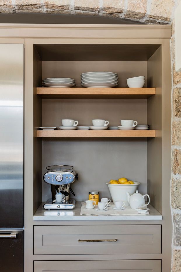 Close-up of a built-in kitchen coffee station set within grey-beige cabinetry. Open shelves display stacked white crockery and cups, while the lower stone benchtop holds an espresso machine, teapot, milk jug, and a bowl of lemons. A stainless-steel refrigerator is partially visible at left, and a rustic stone wall surrounds the joinery.
