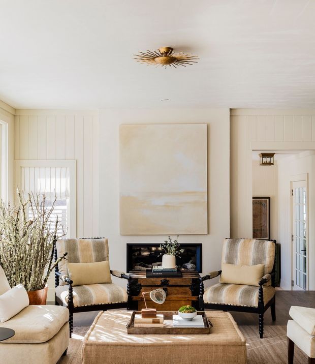 Bright, refined living room with a fireplace, two upholstered armchairs, a large ottoman-style coffee table, and a neutral abstract artwork. White vertical wall panelling, warm timber flooring, and a decorative brass ceiling light create a classic coastal-inspired interior.