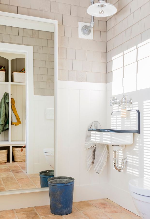Bright, compact bathroom with pale rectangular wall tiles, white vertical wall panelling, terracotta-toned tiled flooring, and a vintage-style wall-mounted basin with exposed chrome plumbing. A large mirror reflects an adjacent toilet and open storage area. Strong sunlight enters through blinds, creating striped shadows across the basin and wall.