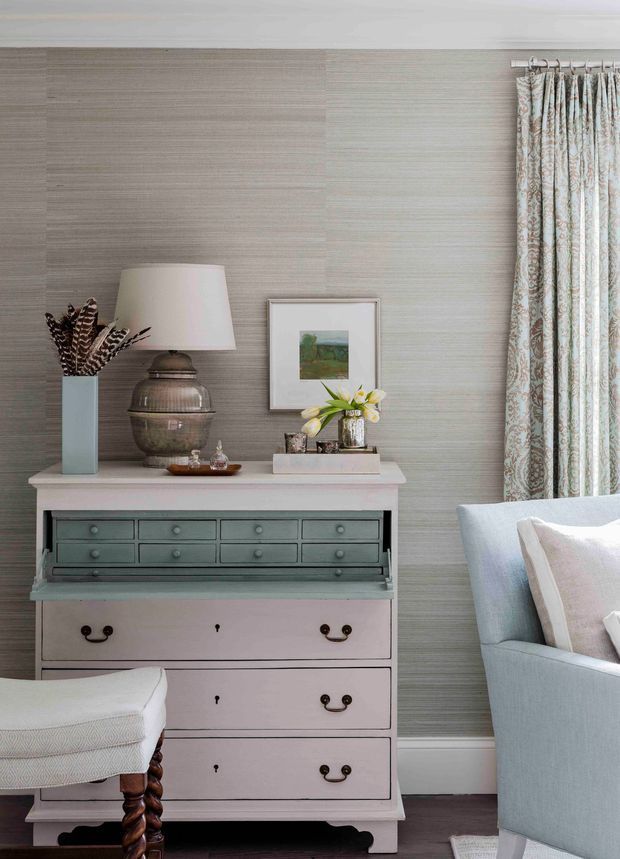 A refined traditional interior vignette featuring a painted multi-drawer chest with a grey-green upper section and pale lower drawers, styled with a table lamp, decorative vase, framed artwork and flowers. Textured taupe-grey wallcovering, white trim, patterned curtains and light upholstered seating create a soft, classic layered look.