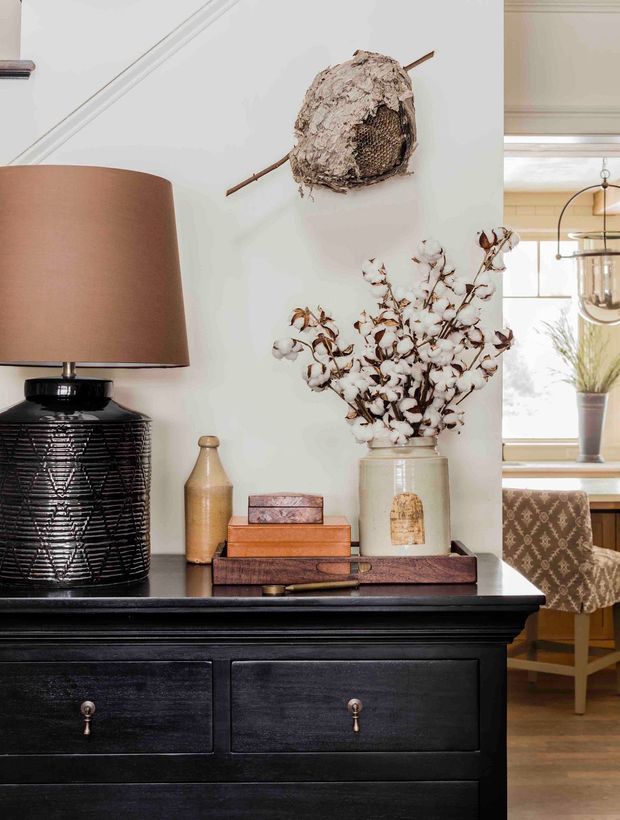 A styled interior vignette featuring a black painted timber console or sideboard with drawers, a ceramic table lamp, stacked books, a vase of cotton stems, and a rustic woven wall object. Through the opening, a bright dining area with a timber table, upholstered chairs, pendant lighting, and a window is visible.