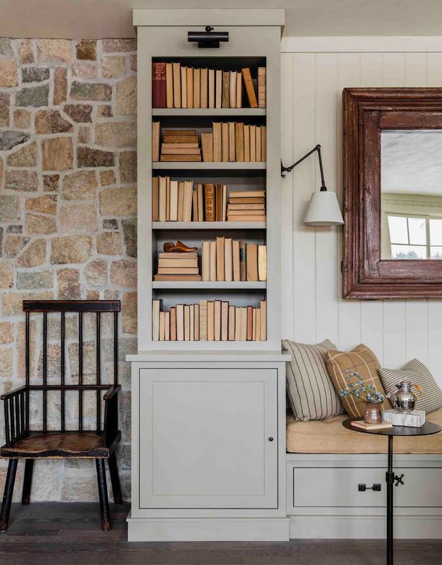 A cozy traditional sitting area featuring a built-in grey bookcase filled with books, set between a rough stone fireplace wall and white vertical wall panelling. A timber-framed mirror, wall-mounted reading lamp, built-in window seat with neutral cushions, black metal side table, and dark timber flooring complete the warm, layered interior.