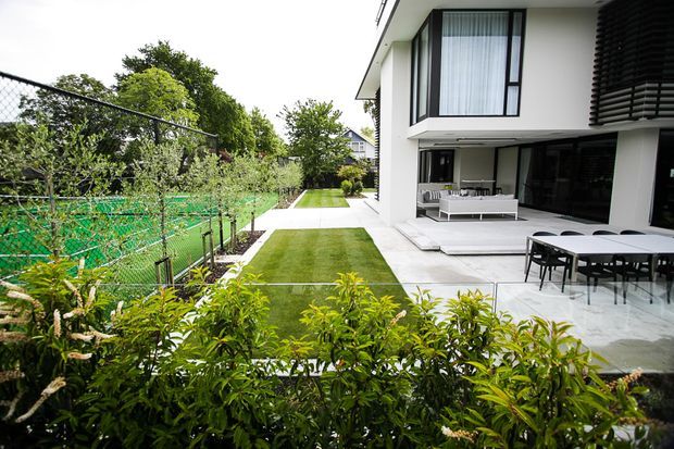 Contemporary two-storey residence viewed from a landscaped side garden, with clean white rendered walls, extensive black-framed glazing, a covered outdoor living area, and a paved dining terrace beside a manicured lawn. A fenced green sports court and hedging sit along the property boundary.