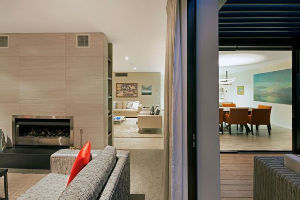 Interior view of a contemporary open-plan home, showing a living room with a large pale timber-clad fireplace wall and built-in firebox, upholstered seating, and a connected lounge area. Through wide glazing and an open doorway is a dining room with a timber dining table, orange-brown chairs, artwork, and a covered timber deck or terrace with dark overhead slatted screening.