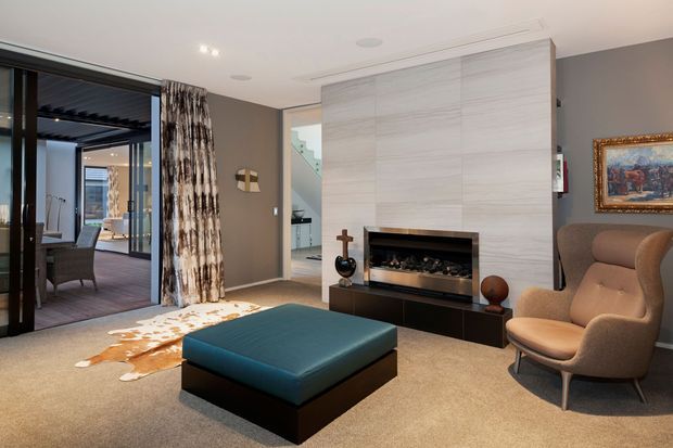 A refined contemporary living room with a large pale stone feature wall and built-in fireplace, low dark joinery, a deep teal ottoman, and a sculptural upholstered lounge chair. Glazed doors open to an adjoining dining or outdoor area, while patterned curtains, artwork, and warm neutral finishes add visual richness.