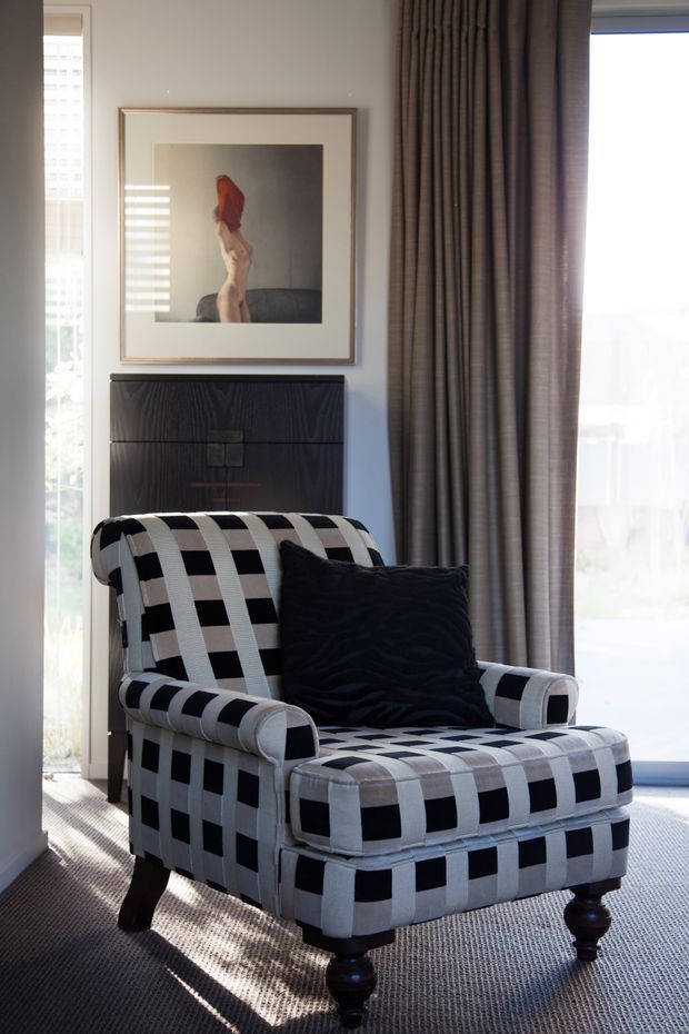 A refined sitting area featuring a bold black-and-white checked upholstered armchair with matching footstool, a dark wood console or cabinet behind it, heavy floor-length taupe curtains, framed artwork, and carpeted flooring. Strong daylight enters through tall windows or glazed doors, creating a monochrome, tailored interior.