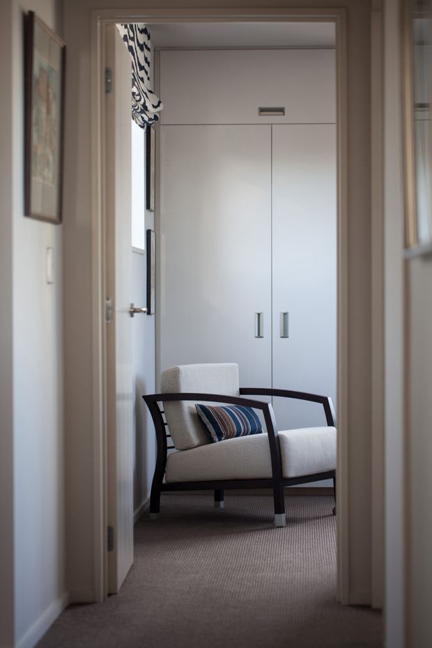 A view through a doorway into a compact, softly furnished sitting room or bedroom lounge. The space features a dark wood-framed upholstered armchair with a striped cushion, pale built-in cabinetry with simple metal pulls, and wall-to-wall neutral carpet. A patterned Roman blind and framed artwork add restrained decorative detail. The palette is warm and muted, with classic trim and understated furnishings.