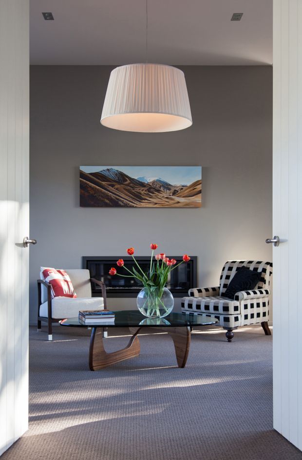 A refined living room viewed through open double doors, featuring a grey painted wall, grey carpet, a central black fireplace, a large pleated pendant light, landscape artwork, a glass-topped sculptural timber coffee table, and two contrasting lounge chairs. Natural light creates a calm, curated atmosphere.