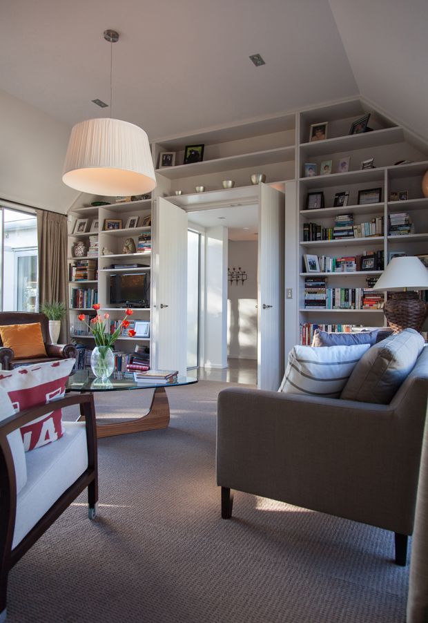 A warm, refined living room with built-in white shelving filled with books, framed photographs and decorative objects. Two upholstered armchairs and a sofa surround a glass-topped coffee table, with a large pleated pendant light overhead. A doorway opens to a hallway, while tall windows provide natural light from the left.