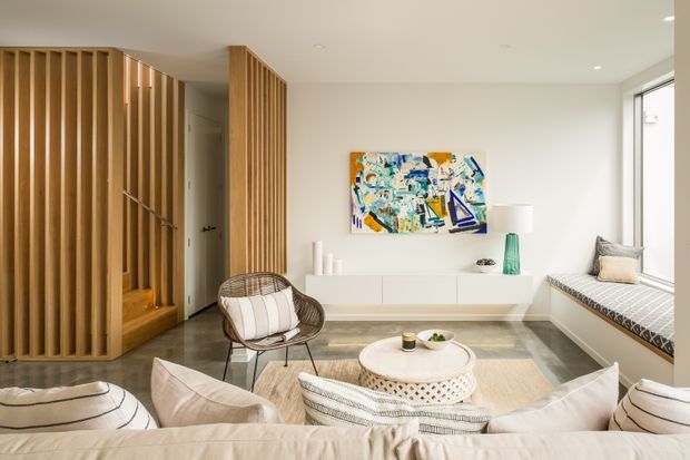 Bright, refined living room viewed from behind a large sofa, featuring a built-in window seat, low white media cabinetry, a round coffee table, neutral upholstered furnishings, and a colourful abstract artwork. A vertical timber slatted screen encloses the stairway and partially separates the hallway from the living area. The space has recessed lighting, pale walls, and large glazing.