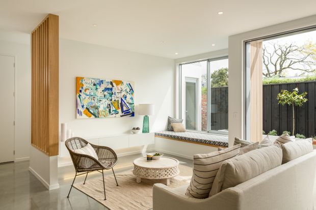 Bright contemporary living room with a cream sectional sofa, sculptural round coffee table, woven lounge chair, built-in white cabinetry, colourful abstract artwork, and large windows opening toward a private courtyard. A vertical timber slat screen defines the entry edge of the room, while a built-in window seat adds seating.