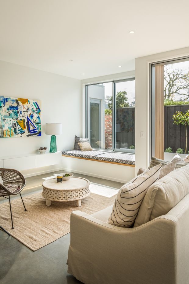 Bright, compact contemporary living room with a large cream slipcovered sofa, patterned neutral cushions, woven rug, round white coffee table, and a sculptural lounge chair. Full-height windows and glazing bring in abundant daylight and overlook a private courtyard. A colourful abstract artwork and turquoise table lamp provide accents, while a built-in white media/storage unit keeps the room streamlined.