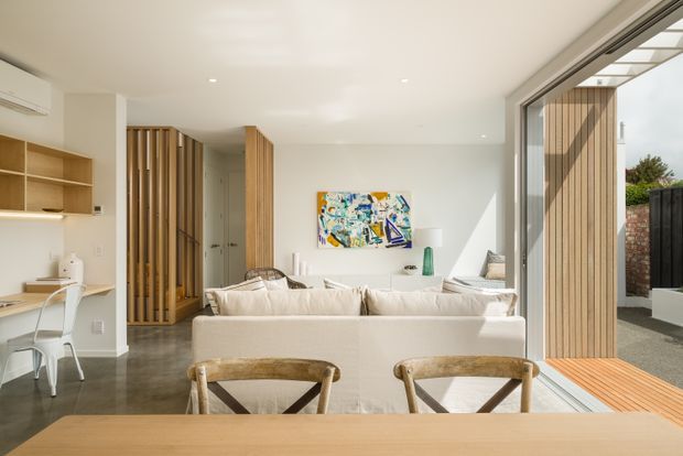 Bright, open-plan contemporary living and dining area with a cream sectional sofa, timber dining table and chairs, built-in timber shelving, vertical timber screening, polished concrete flooring, and large sliding glazed doors opening to an outdoor deck or courtyard. Neutral finishes are complemented by colourful abstract artwork and strong natural light.