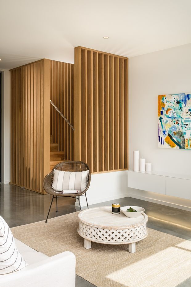 Bright contemporary living room with a white built-in floating media console, large pale rug, round woven coffee table, and a sculptural lounge chair. A striking timber slatted screen forms a feature wall and partially conceals a stairway, while an abstract colourful artwork adds an accent on the white wall. The room has warm natural light, polished concrete flooring, recessed ceiling lighting, and a clean open-plan feel.