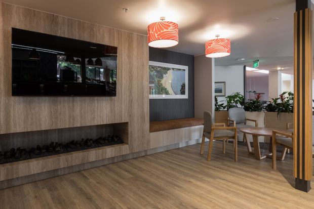 A warmly lit contemporary interior lounge or reception area with timber-look flooring, a full-height wood-grain feature wall, wall-mounted television, long linear fireplace, built-in bench seating, and a small dining or waiting area. Red-and-white pendant lights, timber-framed chairs, plants, and glazed partitions add colour and definition. A dark vertical timber screen frames the right side of the space.
