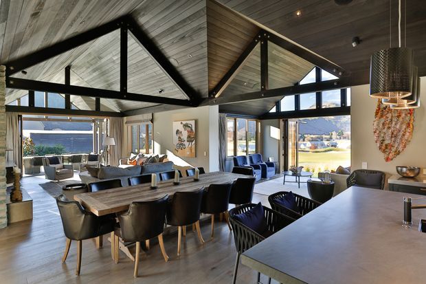 A spacious open-plan contemporary living, dining and kitchen area with dramatic exposed timber cathedral ceilings and black triangular structural framing. Floor-to-ceiling glazing provides expansive views, while a large timber dining table, upholstered dining chairs, lounge seating and a substantial kitchen island create a refined, warm interior.