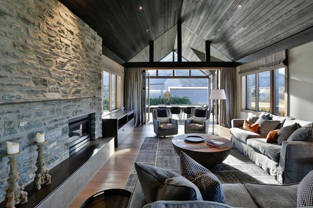 A spacious, refined living room with a dramatic vaulted dark timber ceiling, exposed black structural beams, extensive glazing, and a large rough-stone fireplace wall with integrated dark joinery. The room is furnished with a large sectional sofa, lounge chairs, layered rugs, and a substantial round timber coffee table, with views toward an outdoor terrace or garden beyond.