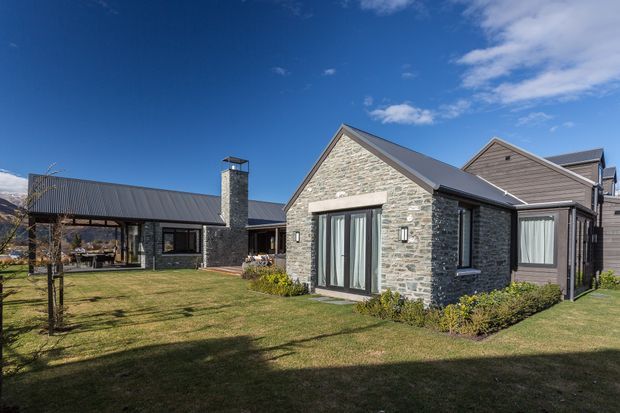 Exterior view of a low-rise residential building arranged around a broad landscaped lawn. The main dwelling features a gabled volume clad in grey natural stone with dark metal roofing, large glazed doors, and adjoining dark horizontal timber-clad sections. A covered link or wing and stone chimney are visible to the left, with mountain terrain in the distant background.