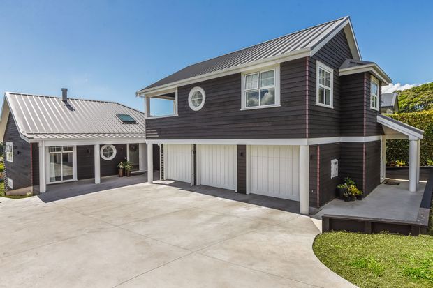 Exterior view of a multi-level residential building with dark horizontal cladding, light corrugated metal gable roofs, white-framed windows, multiple garage doors, and a broad concrete driveway. The design includes a covered upper-level balcony or walkway and a sheltered side parking area.