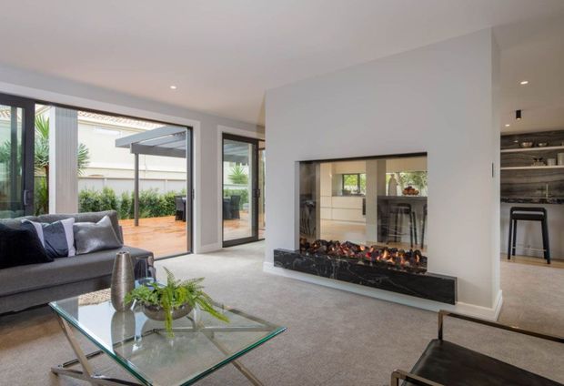 A bright contemporary living room with a large grey sectional sofa, glass coffee table, and a wide built-in gas fireplace set into a white feature wall. Full-height glazed doors open to a paved outdoor terrace and garden, while an opening at right reveals a compact kitchen or bar area with open shelving and stools. Neutral finishes and abundant glazing create an airy, connected indoor-outdoor space.