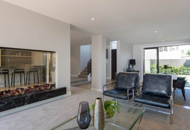 Contemporary open-plan living room with a glass-fronted black marble fireplace, two black leather lounge chairs, a glass coffee table, and neutral carpeting. A dining or kitchen area is visible behind the fireplace, with stairs and large glazed doors opening toward a planted outdoor area.