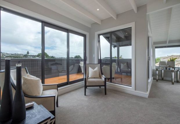 Bright, refined sitting area with two upholstered armchairs beside expansive dark-framed glazing and a mirrored wall panel. The room features grey carpet, light grey walls, white painted exposed ceiling beams, and views onto a timber deck and covered outdoor dining area. The restrained palette and strong indoor-outdoor connection create an airy contemporary coastal feel.