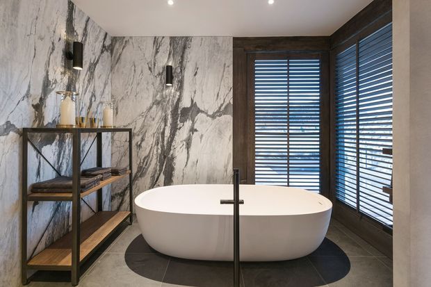 A sophisticated contemporary bathroom featuring a freestanding white oval bathtub, dark floor tiles, dramatic marble-look wall cladding, dark timber window frames with horizontal blinds, and a black metal open shelving unit. Recessed ceiling lights and black wall sconces provide focused illumination, creating a refined, high-contrast interior.