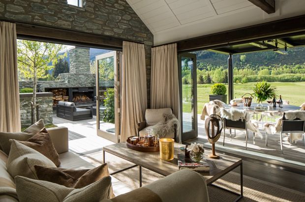 A refined, light-filled living room with large floor-to-ceiling glazing opening onto a covered outdoor dining terrace and landscaped grounds. The room features a stone-clad fireplace wall, exposed dark timber beams, neutral upholstered seating, full-height curtains, and a glass-topped coffee table. A substantial outdoor stone fireplace and seating area are visible beyond the open glazing.