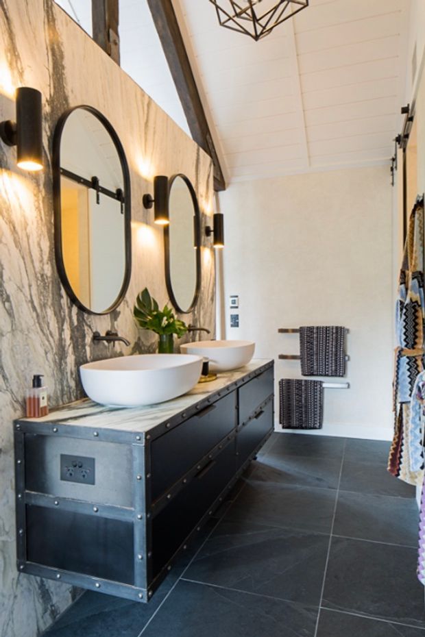 A stylish bathroom featuring a long industrial-style double vanity with two white vessel basins, dark metal cabinetry with riveted detailing, and a dramatic marble-look wall finish. Arched black-framed mirrors and matching wall sconces line the vanity, while dark large-format floor tiles, exposed timber ceiling beams, white panelled ceiling boards, and woven wall-mounted towel rails complete the space.