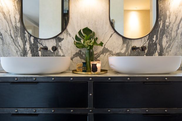 A sophisticated bathroom vanity with two white vessel basins set on a dark, industrial-style double vanity. The wall is clad in dramatic grey-and-white veined marble, complemented by two rounded black-framed mirrors, wall-mounted dark fixtures, and warm decorative lighting and accessories.