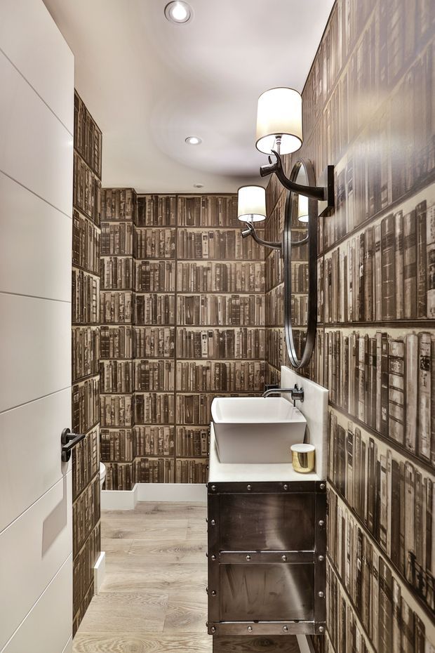 A narrow contemporary bathroom viewed from the doorway, featuring a wall-mounted white vessel basin on a dark metal vanity, a large round mirror, twin wall sconces, pale wood-look flooring, and dramatic full-height wallpaper depicting rows of antique books. The left wall and door are finished in plain white panels.