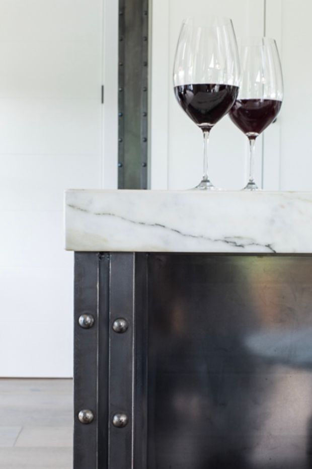 Close-up architectural detail of a dark metal kitchen or bar island with exposed riveted vertical supports and a thick white veined marble benchtop. Two red wine glasses sit on the surface. The surrounding interior is bright and minimal, with white wall cabinetry or paneling visible in the background.
