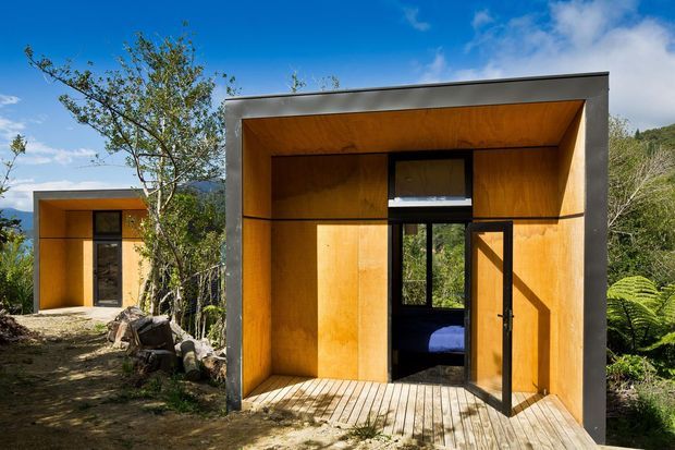 Exterior view of two compact modern cabin-like structures set in a wooded, mountainous landscape. The foreground structure has a simple rectangular form with a deep recessed entrance, warm timber-lined walls and ceiling, dark grey structural framing, a timber decking threshold, and an open glazed door revealing a small bedroom inside. A second matching unit is visible in the background.
