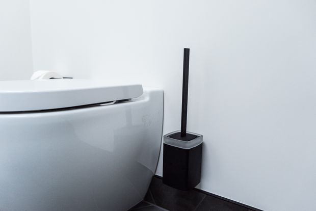 Close-up of a contemporary white toilet beside a black toilet brush holder in a minimalist bathroom. The room has smooth white walls, dark tiled flooring, and a clean, high-contrast monochrome palette.