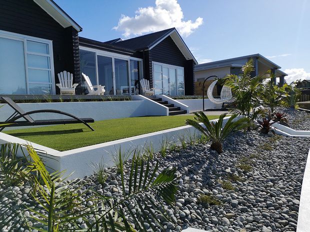 Exterior view of a contemporary single-storey residential home with dark horizontal cladding, steep gabled roof forms and extensive glazed outdoor living areas. The landscaped front yard features raised white rendered garden beds, synthetic lawn, dark river stones, palms and succulents, with outdoor lounge furniture and a hanging egg chair.